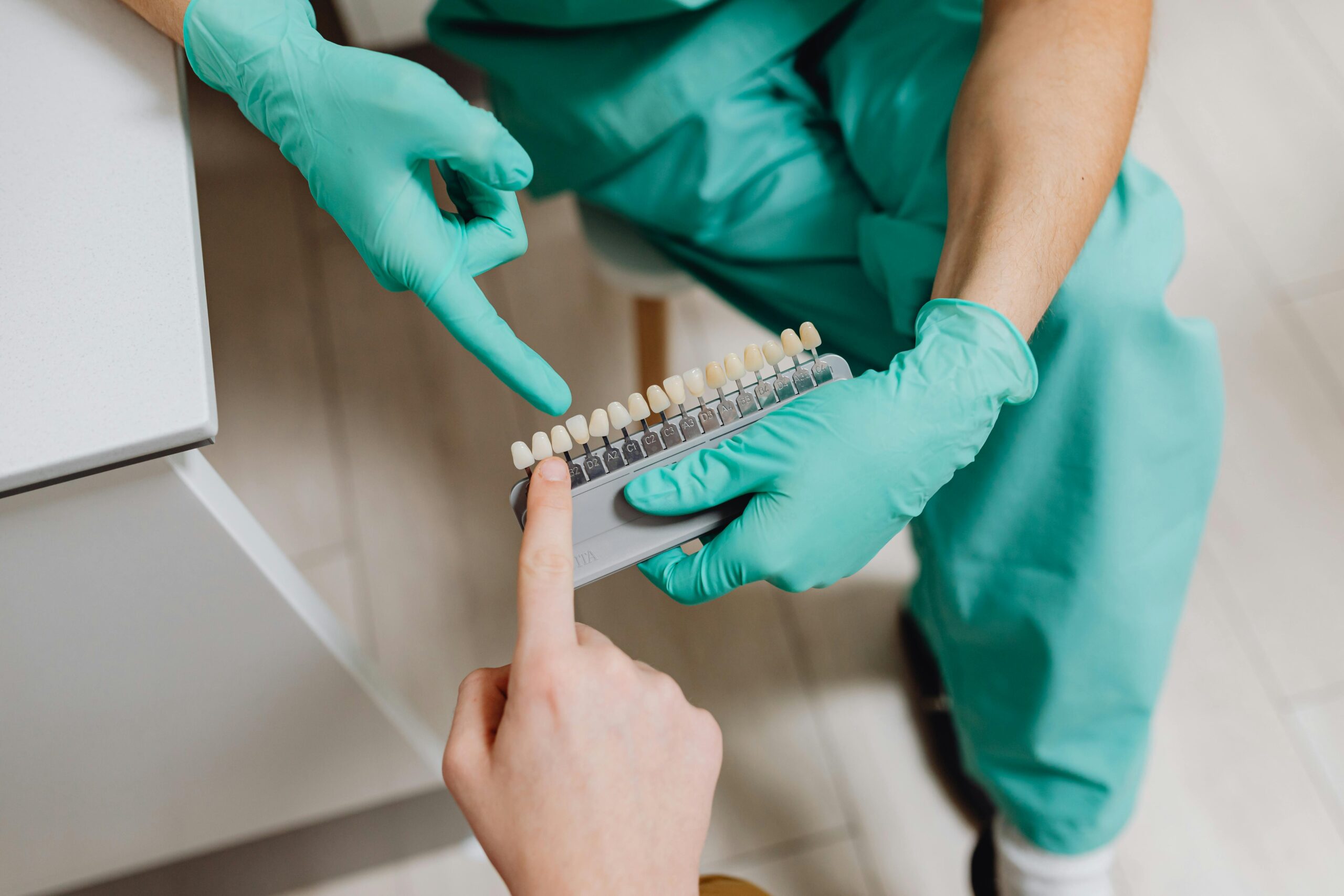 A dental professional in gloves holding a teeth shade guide while assisting a patient indoors.